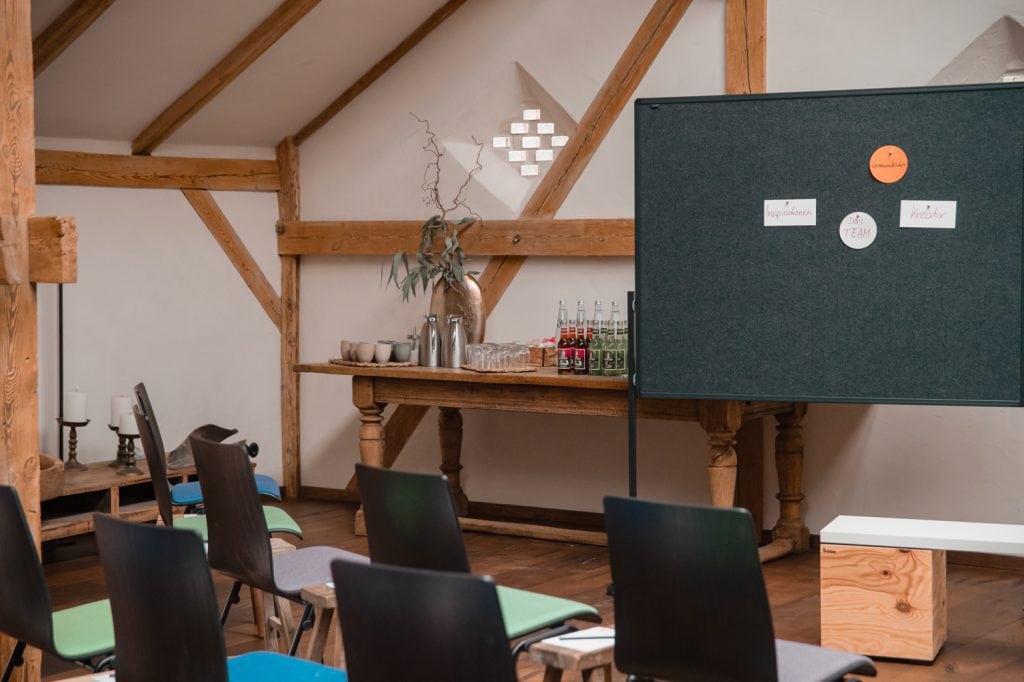 Entspannter Seminarraum mit Holzbalken, modernen Sitzgelegenheiten und Pinnwand f&uuml;r kreative Workshops.