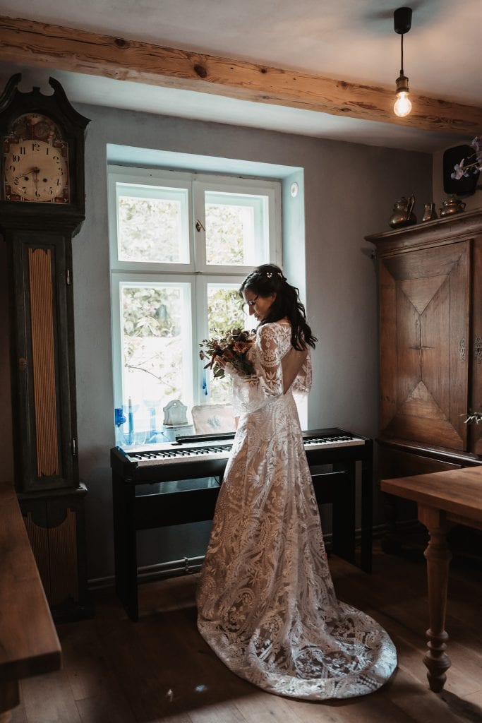 Elegant Braut im Hochzeitskleid mit Blumen vor Fenster, gem&uuml;tliches rustikales Interieur.
