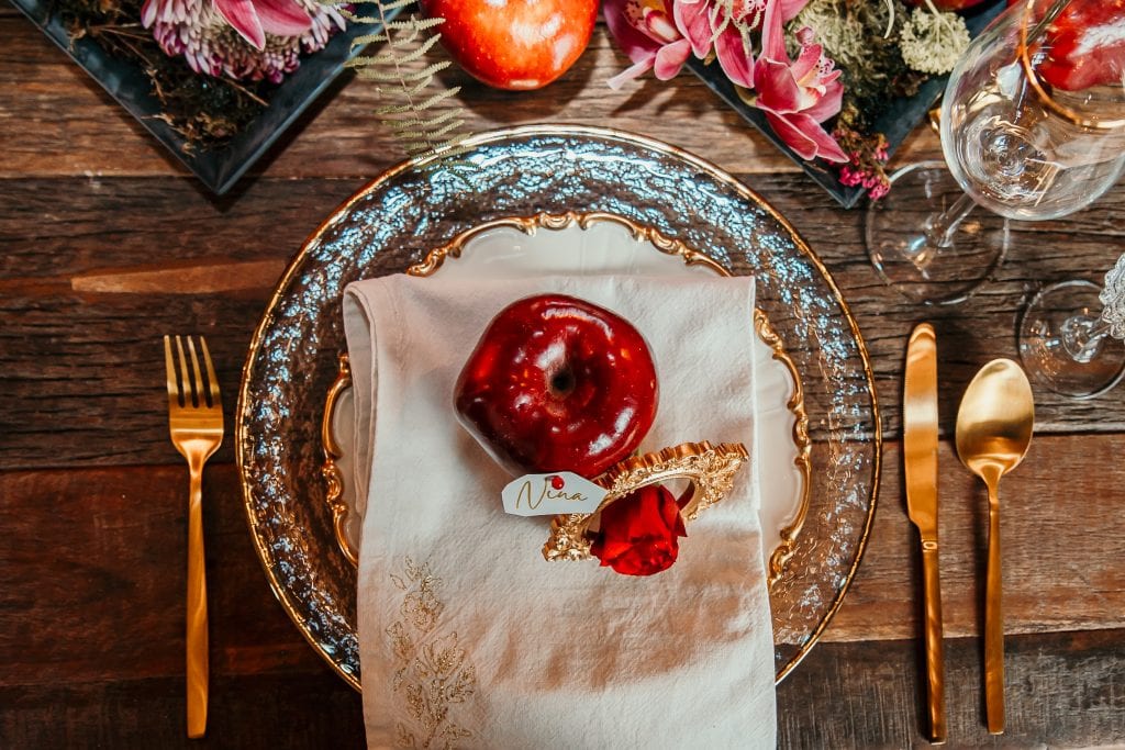 Frühstückstisch mit Apfel, goldene Besteck, elegantem Geschirr und Blumenarrangements.