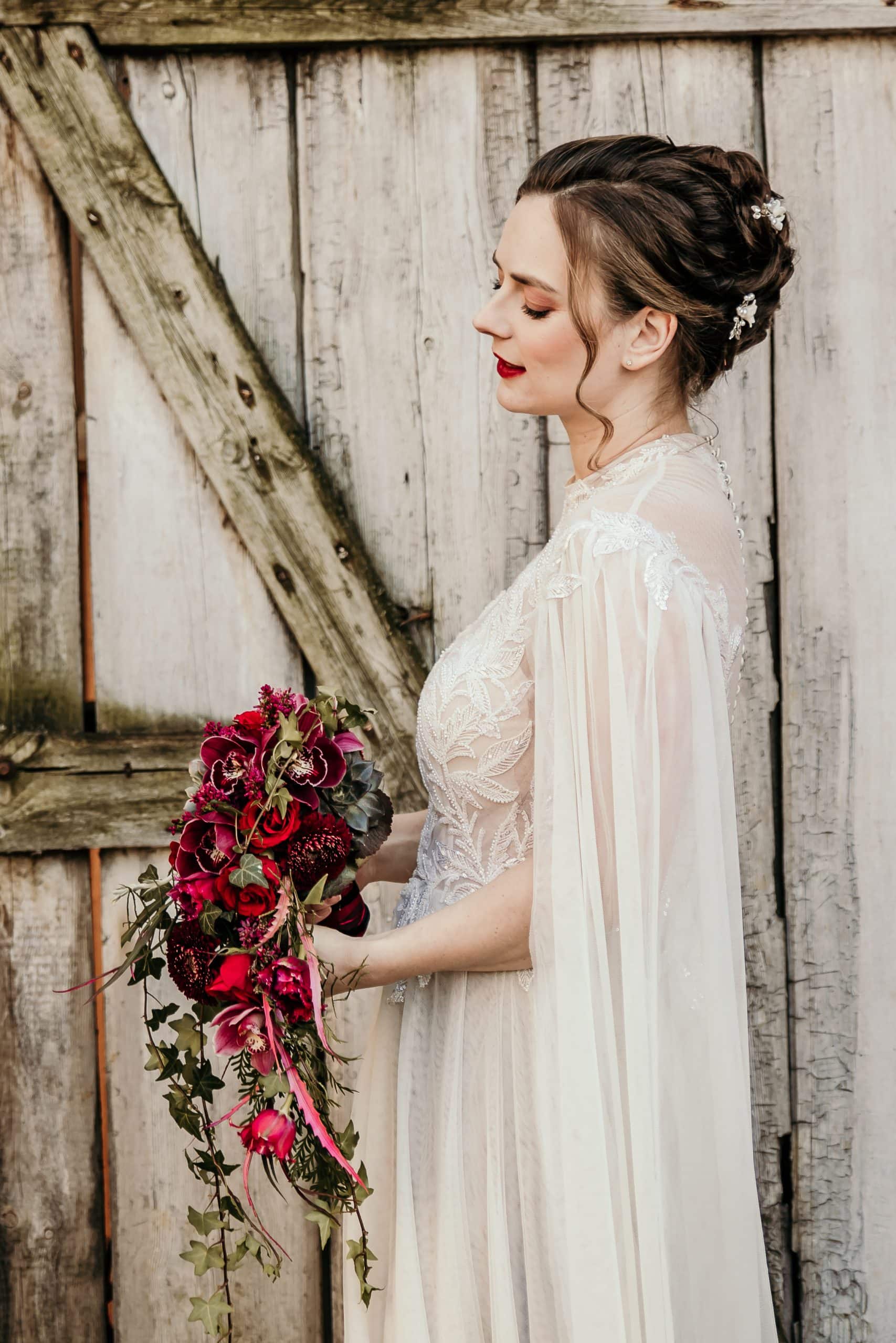 1. Hochzeitsbraut mit rotem Blumenstrauß vor Holzscheune, Naturlook.