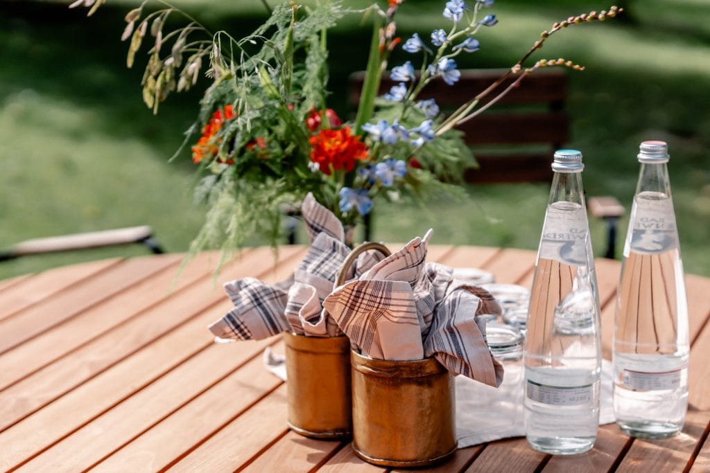 Frühstückstisch im Garten mit Wasserflaschen, Besteck und einer Blumen-Vase für eine gesunde Ernährung.