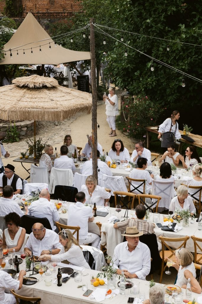 Belebter Garten im stilvollen Caf&eacute; mit zahlreichen G&auml;sten, wei&szlig;er Dekoration und entspannter Atmosph&auml;re.