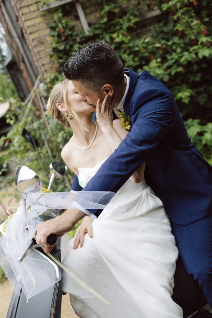 Verliebtes Brautpaar k&uuml;sst sich auf Vespa, romantische Hochzeitsfahrt im Garten, Gut Leben Berlin.