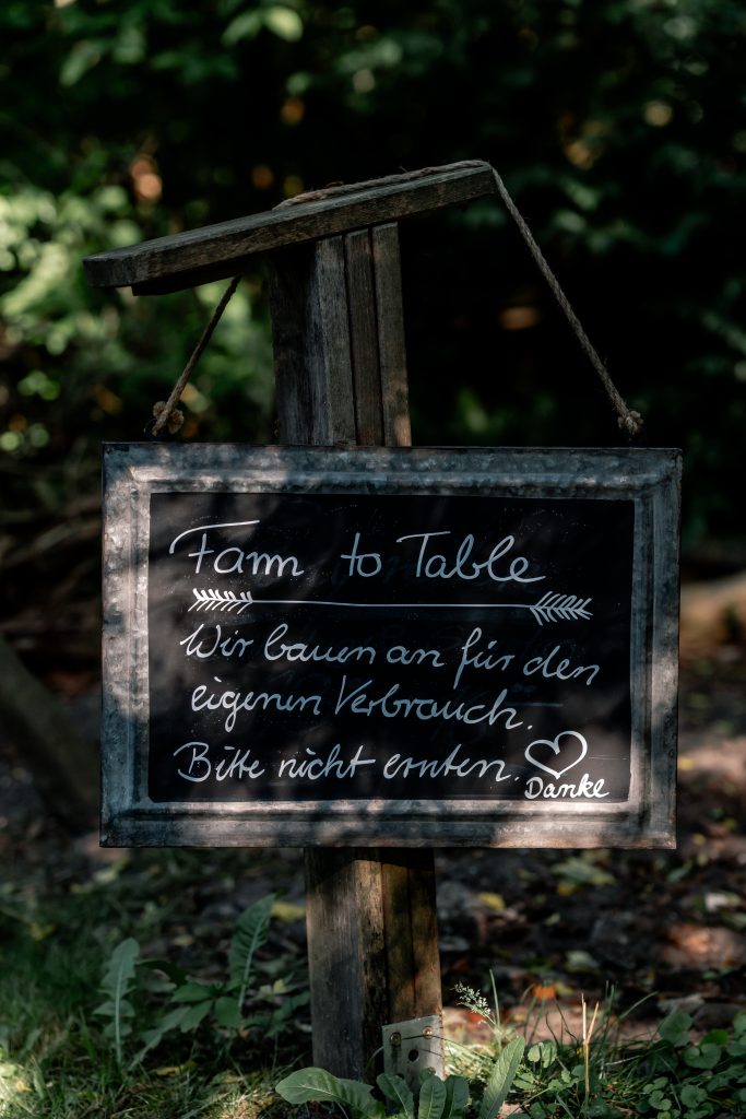 Ein handgeschriebenes Schild mit dem Text "Fam to Table" in einer natürlichen Umgebung, umgeben von Grünpflanzen.