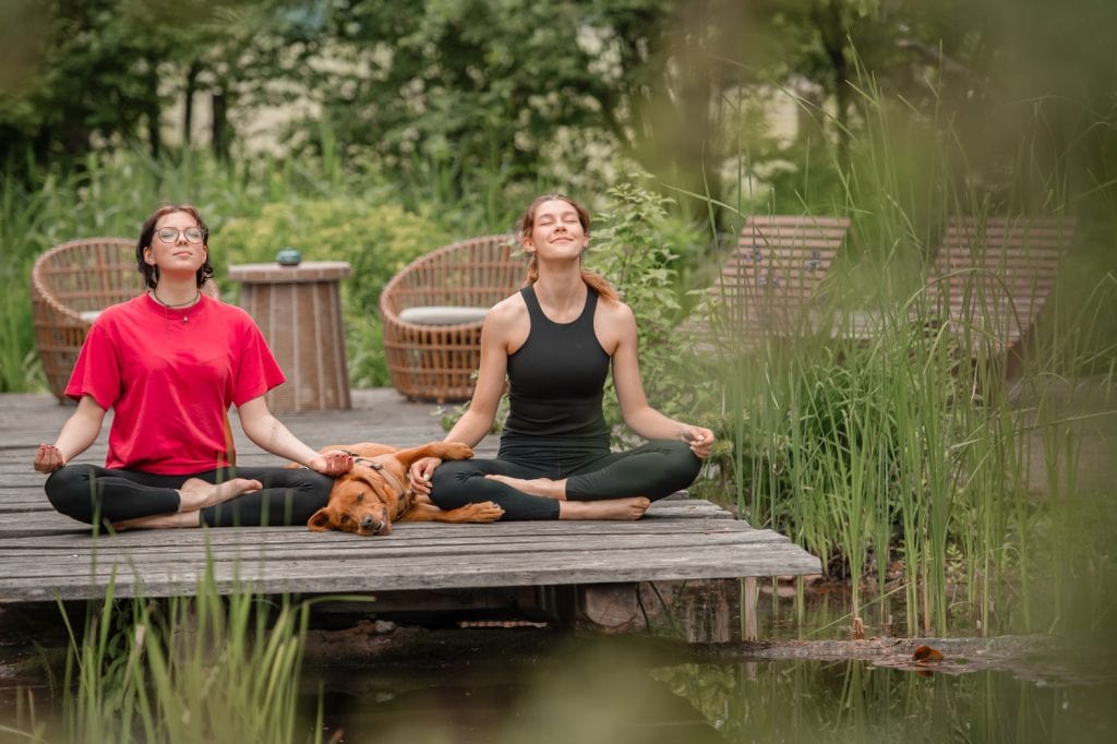 Entspannende Meditation im Garten mit Frau, Frau und Hund an einem ruhigen See, Stressabbau und Achtsamkeitspraxis.