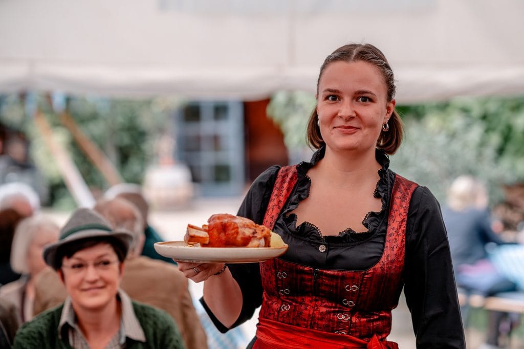 Fr&ouml;hliche Frau in traditionellem Dirndl serviert deutsches Gericht auf festlichem Markt.