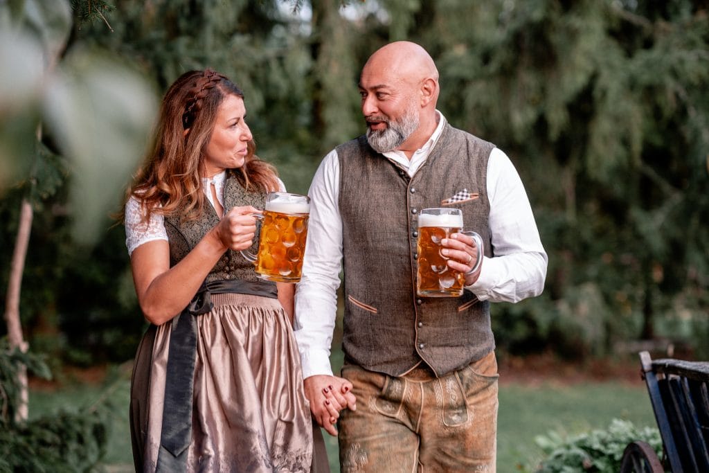 Fröhliches Paar im Dirndl und Lederhose beim Biertrinken im Garten, saisonales Oktoberfest-Feeling.