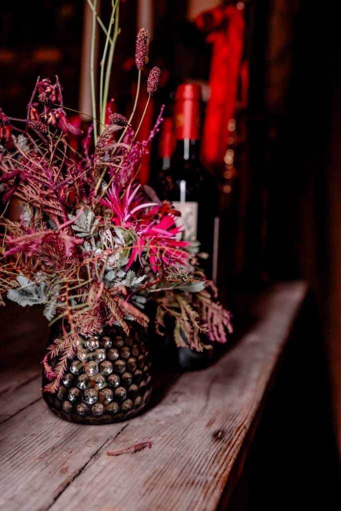 Blumenarrangement in dunklem Vase mit saisonalen Blüten und Blättern, rustikaler Holzuntergrund.