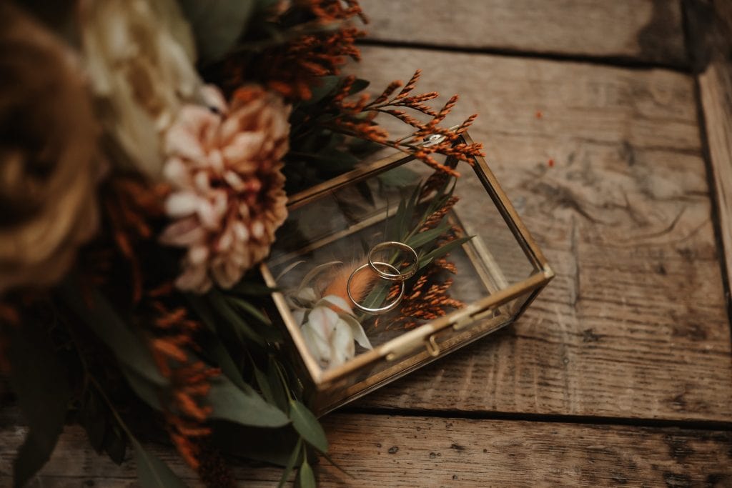 Metallene Ringe in einer Glasbox mit Trockenblumen und Kerze, rustikaler Vintage-Stil.