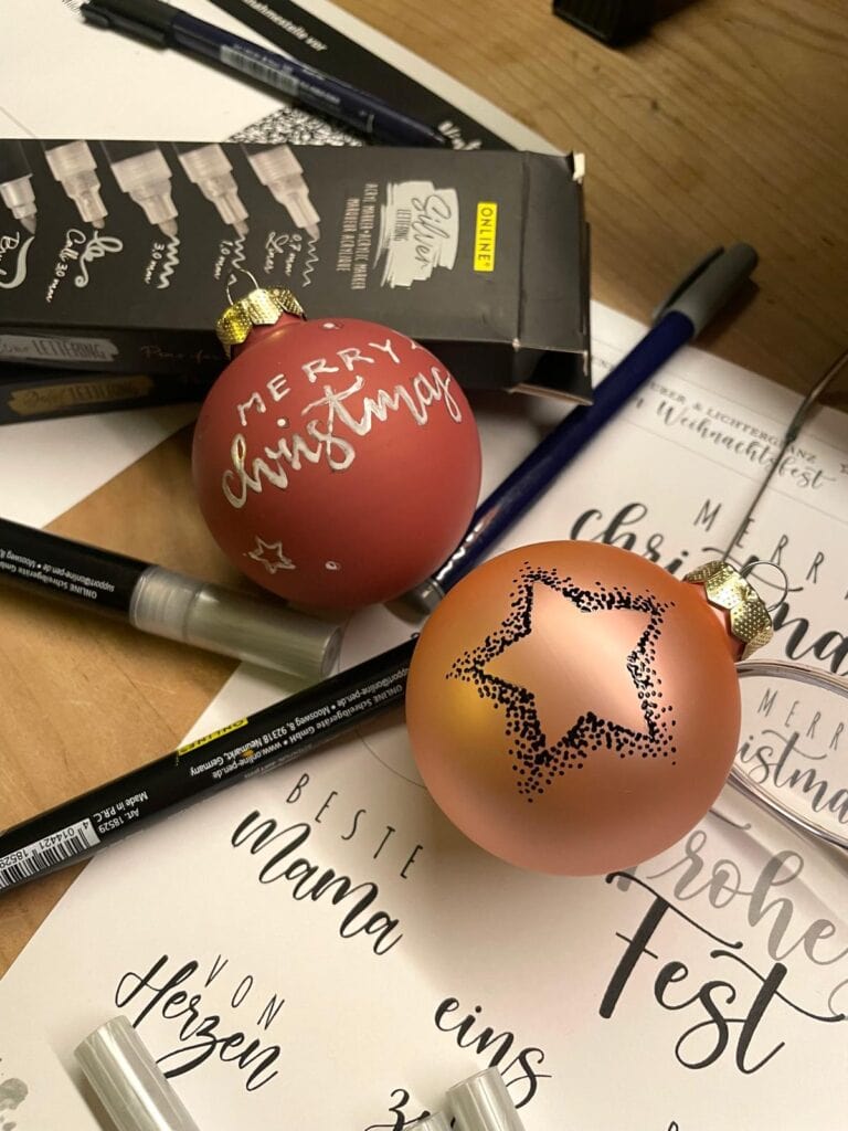 Weihnachtliche Kalligrafie mit Weihnachtskugeln und Schreibutensilien auf Papier.