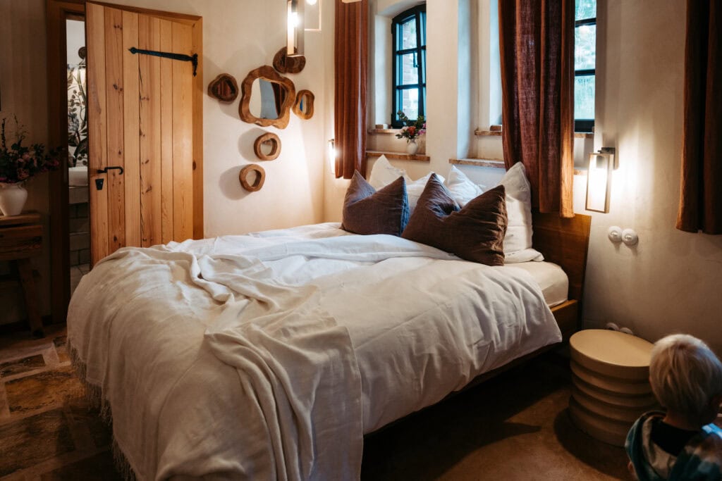Gemütliches Schlafzimmer mit weißem Bett, Holzdeko und Fenster mit roten Vorhängen.